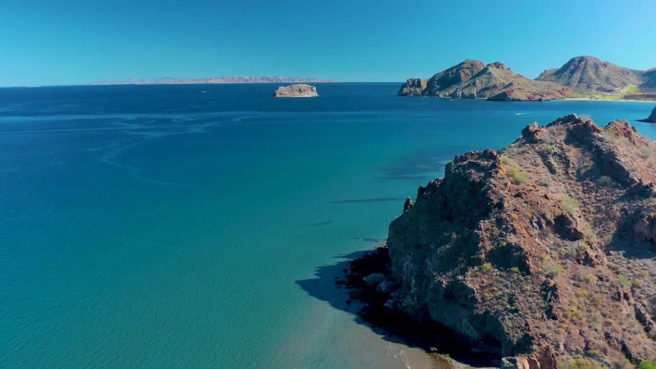 High aerial view in Ligui, Baja California Sur, showing a rugged rocky cliff in the foreground, turquoise coastal water, distant islands, clear blue sky, and no visible people.