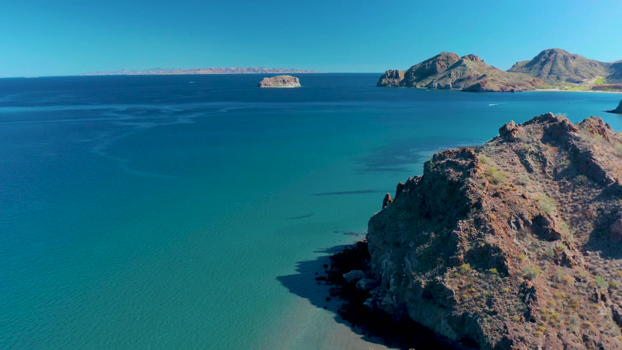 Ligui coastal aerial with a jagged brown outcrop along the lower right, clear turquoise water spreading across the frame, islands on the horizon, and no visible people.