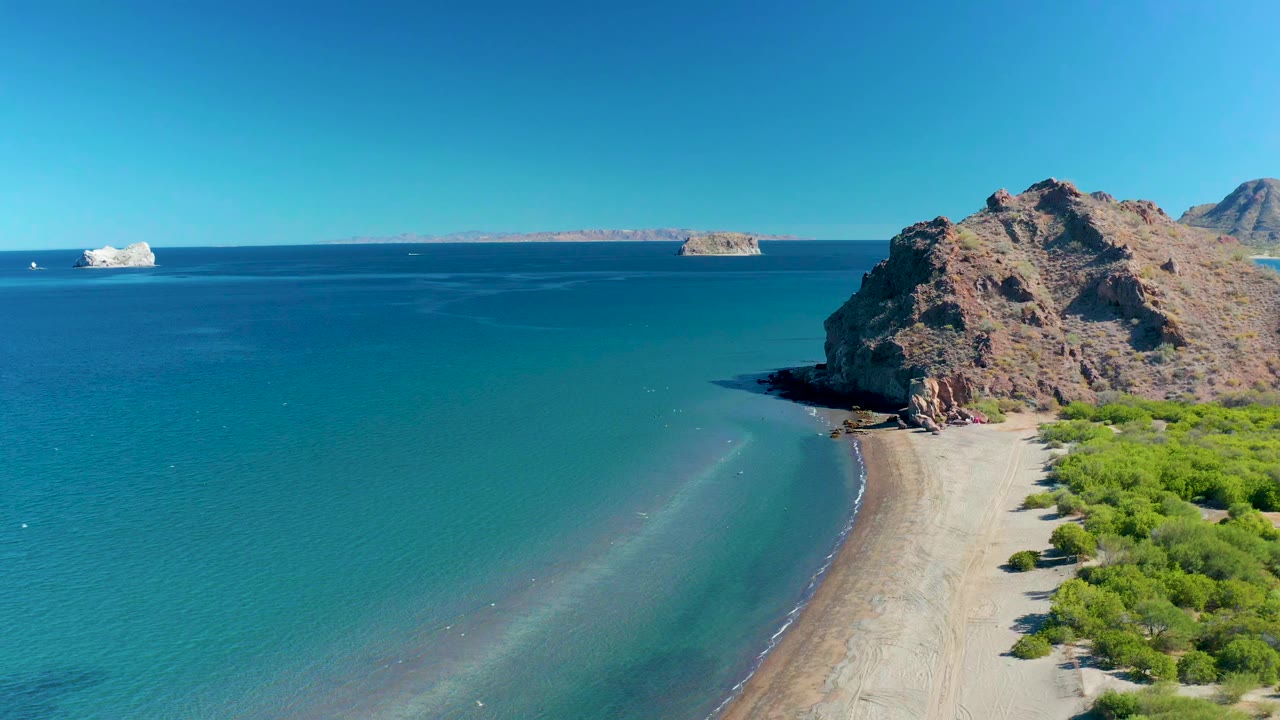 Opening aerial frame over Ligui, Baja California Sur with turquoise water on the left, a pale sandy beach below a rocky outcrop on the right, distant islands on the horizon, and no visible people.