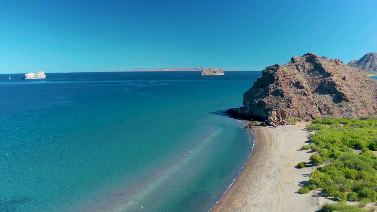 Aerial frame in Ligui, Baja California Sur showing the rocky headland shifted slightly right in the composition, blue-green water spreading across the left side, small wave marks near shore, and no visible people.