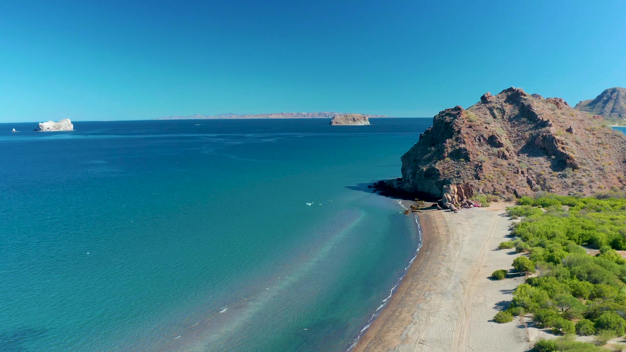 Wide coastal aerial with calm ocean, beach, rocky outcrop, and vegetation.