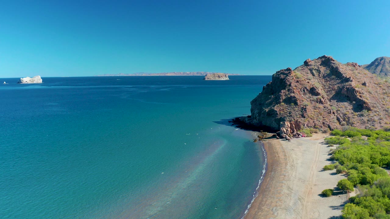 At 0:00, Ligui, Baja California Sur appears from above with turquoise water in the foreground, a sandy beach curving below a rocky cliff, distant islands on the horizon, and no visible people.