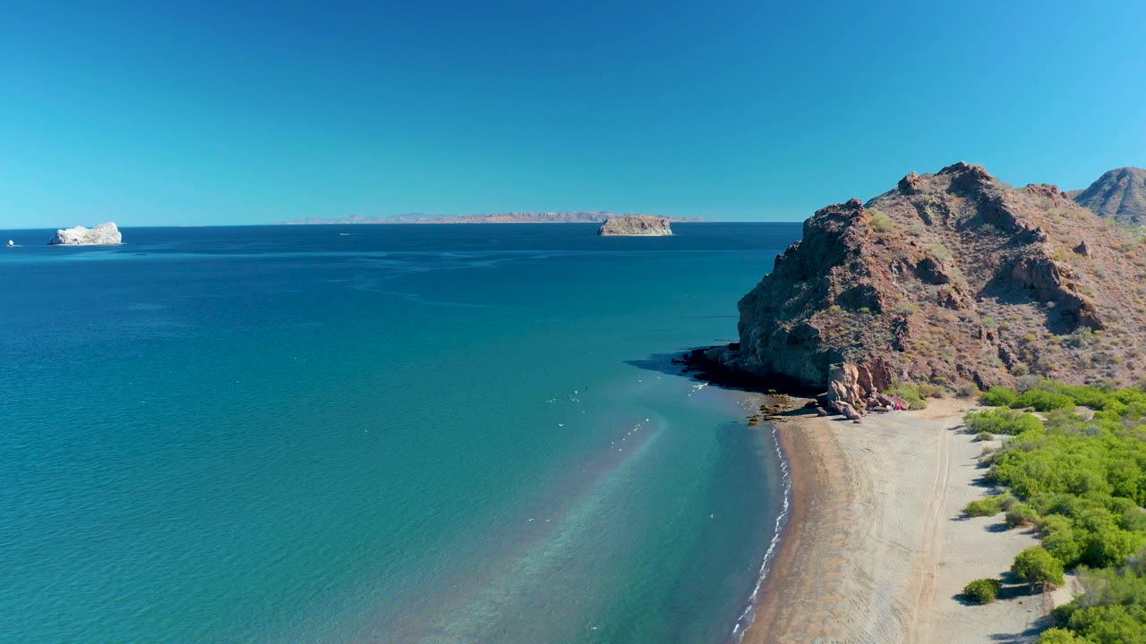 At 0:02, the shallow turquoise water forms a pale diagonal band toward the sandy Ligui beach, while the rocky headland fills the right side and no visible people are present.