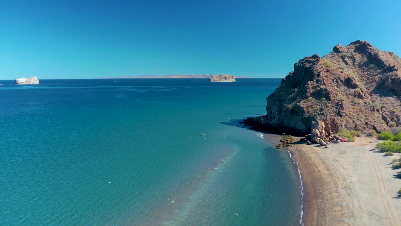At 0:01 in Ligui, Baja California Sur, shallow blue water shows a pale sandbar-like curve near the beach while the rocky headland holds the right edge, with no visible people.