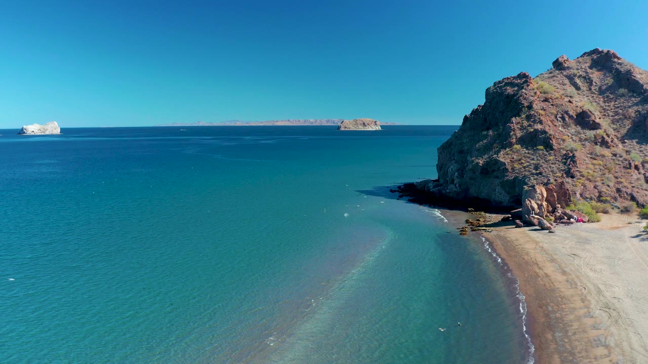At 0:03 in Ligui, Baja California Sur, open turquoise water dominates the left side as small waves reach the sandy beach below the dark shadow of the rocky cliff, with no visible people.
