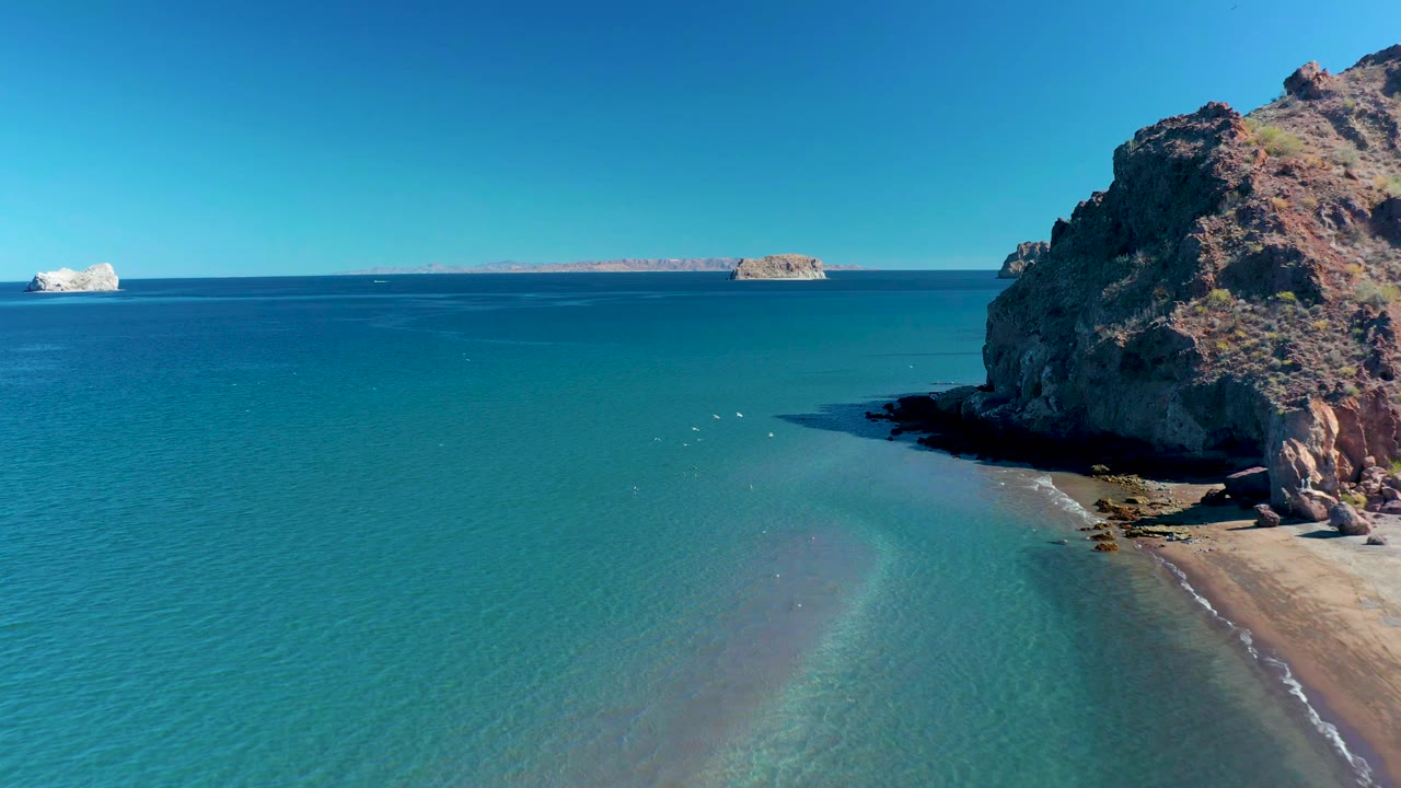 Elevated Ligui coastline frame with open blue water in the foreground, a shaded rocky cliff on the right, faint wave lines over the sandy shallows, distant islands, and no visible people.