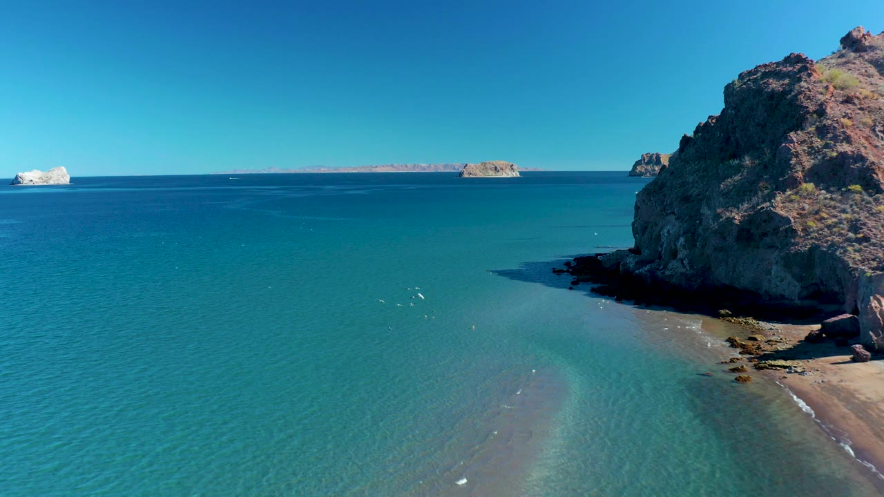 Ligui aerial view showing broad turquoise water in the foreground, a rocky headland on the right, a narrow sandy shore below it, small ripples across the shallows, distant islands, and no visible people.