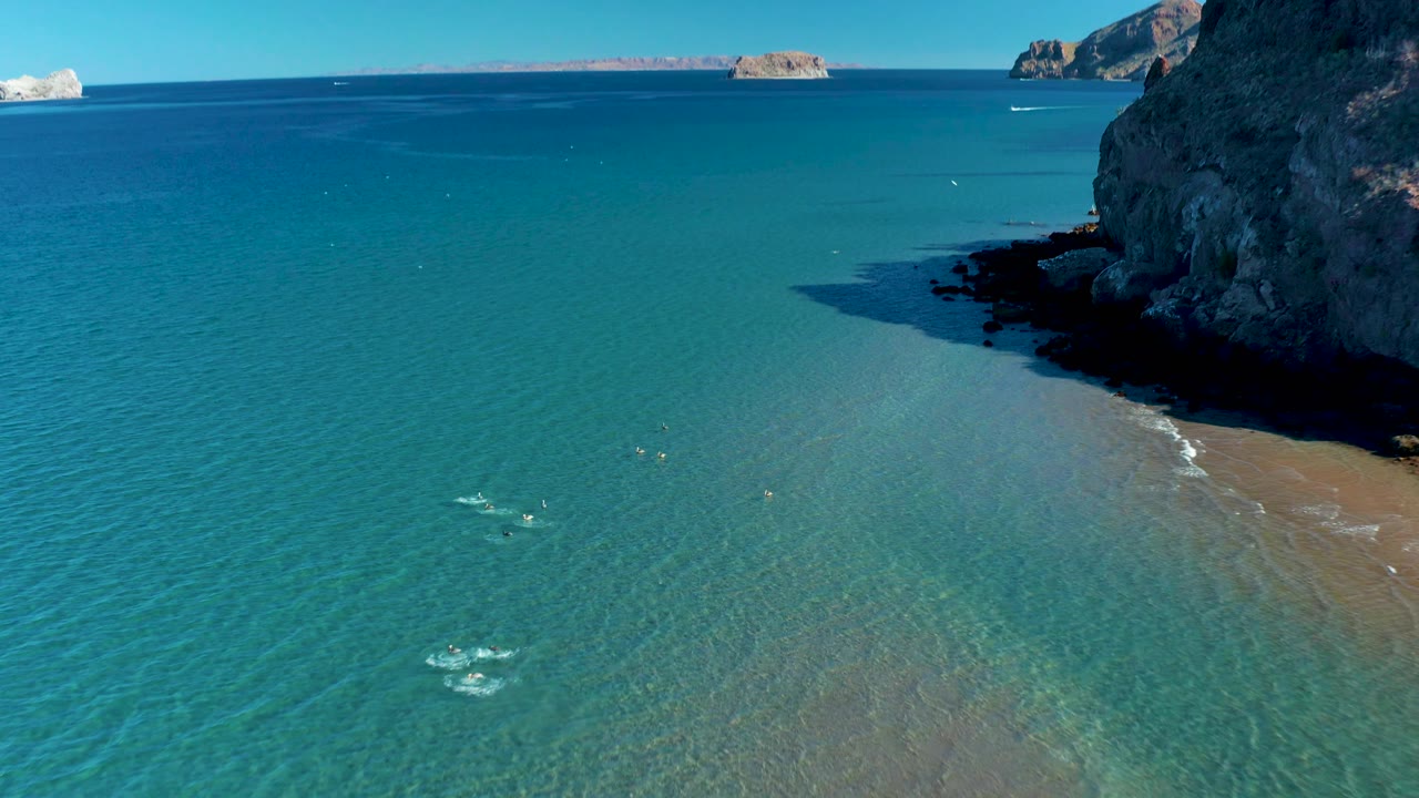 Aerial view of swimmers scattered in the clear shallows below a rocky cliff, with gentle wave lines reaching the sandy beach at right in Ligui.