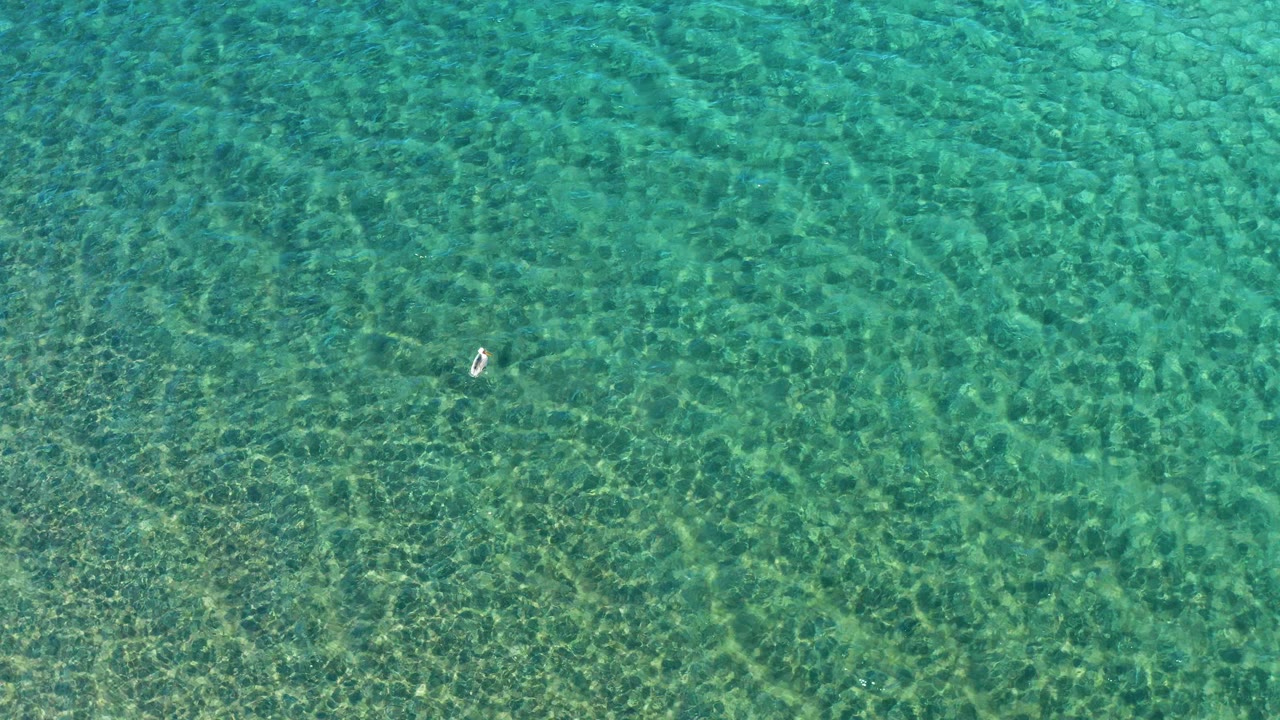 High overhead view of clear turquoise water in Ligui, Baja California Sur, with one small swimmer near the left side and sunlight ripples across the seabed.