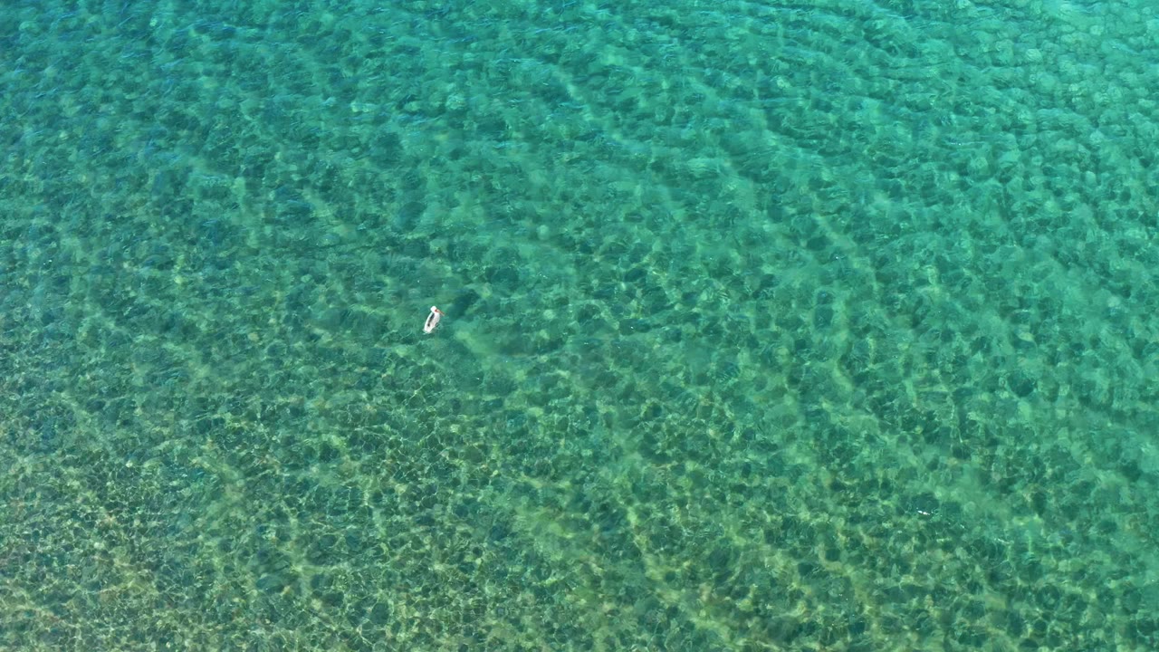 High overhead view in Ligui, Baja California Sur, showing one small swimmer right of center over transparent turquoise water and bright seabed texture.