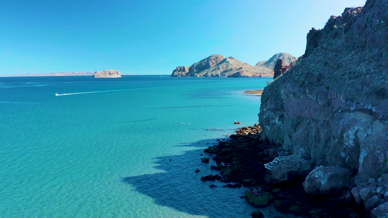 Aerial frame in Ligui, Baja California Sur, Mexico, with the cliff face on the right, clear turquoise water below, a thin white speedboat wake left of center, distant islands, and no visible people.