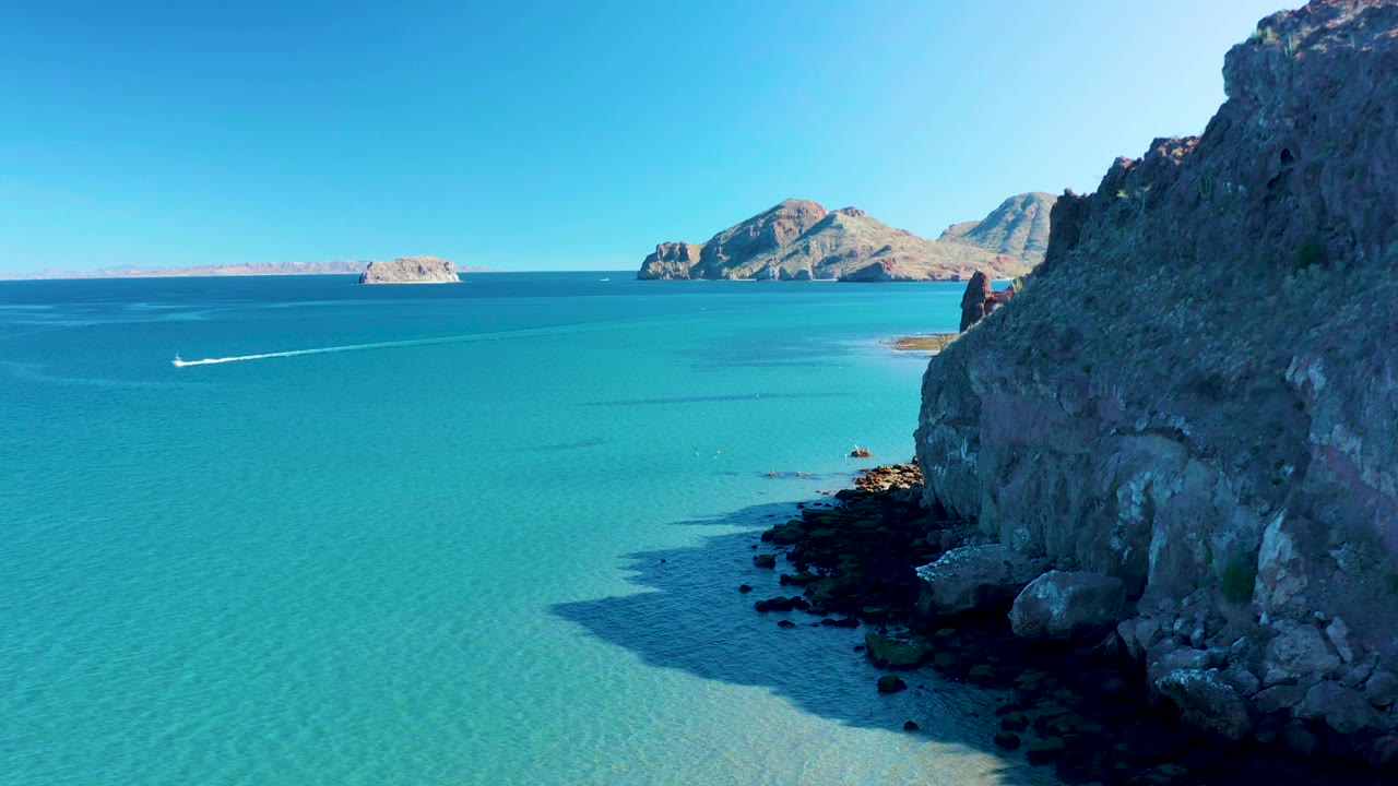 High coastal view in Ligui with the white wake slightly farther across the turquoise sea, the rocky right-hand cliff in shadow, distant islands or headlands, and no visible people.