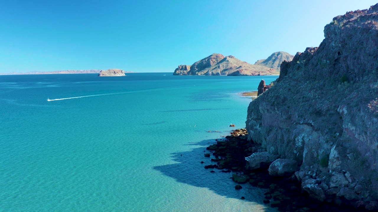 Aerial frame over Ligui coastline showing a broad turquoise water field, the cliff face cropped along the right edge, a boat wake left of center, distant landforms, and no visible people.