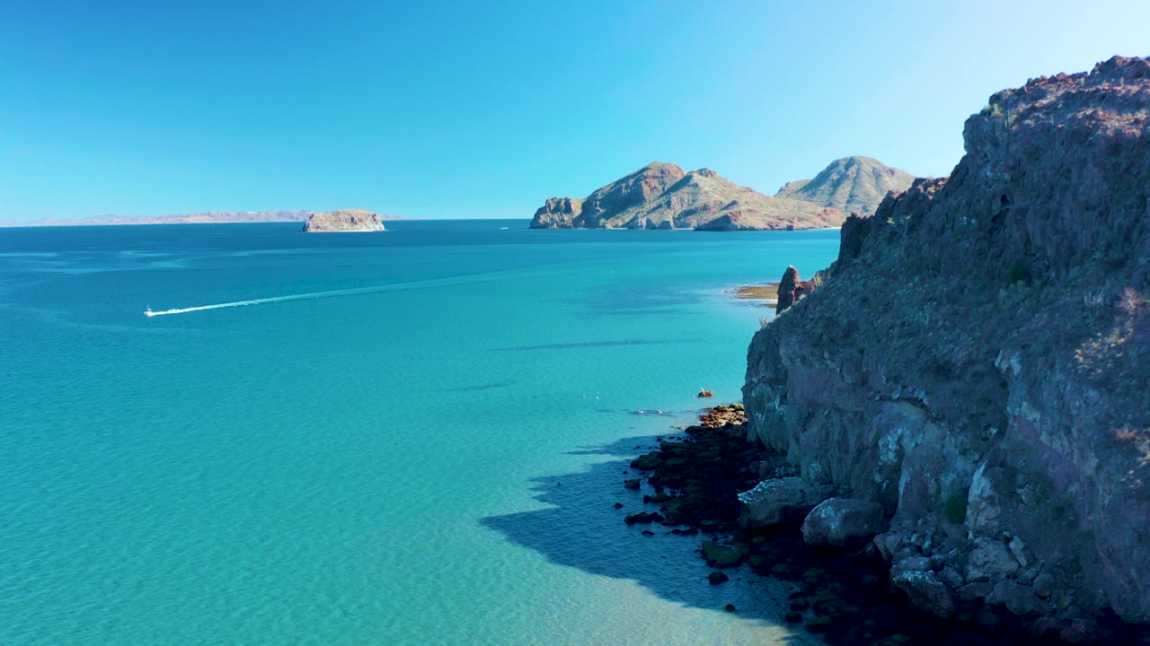 Ligui ocean view with a longer white wake across blue-green water, the rocky cliff occupying the right side, a small sandy shoreline area near the distance, and no visible people.
