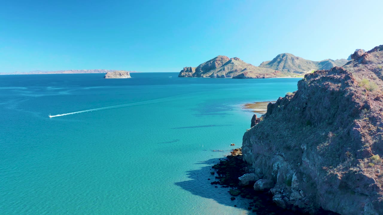 Aerial coastal frame from Ligui, Mexico, showing the boat slightly farther across calm blue-green water, with the rocky right shoreline and low islands under a clear sky, no visible people.