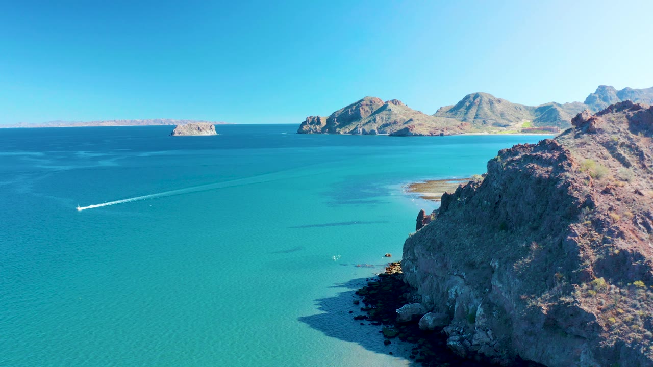 Ligui aerial frame with the boat and wake crossing open turquoise water, a rugged rocky slope filling the right foreground, small sandy shallows near shore, and no visible people.