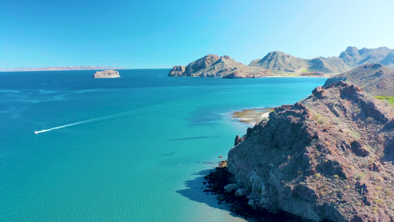 Aerial coastal frame from Ligui showing the boat slightly farther along its route, a long wake across clear turquoise water, rocky foreground cliffs, and blue mountain coastline, no visible people.