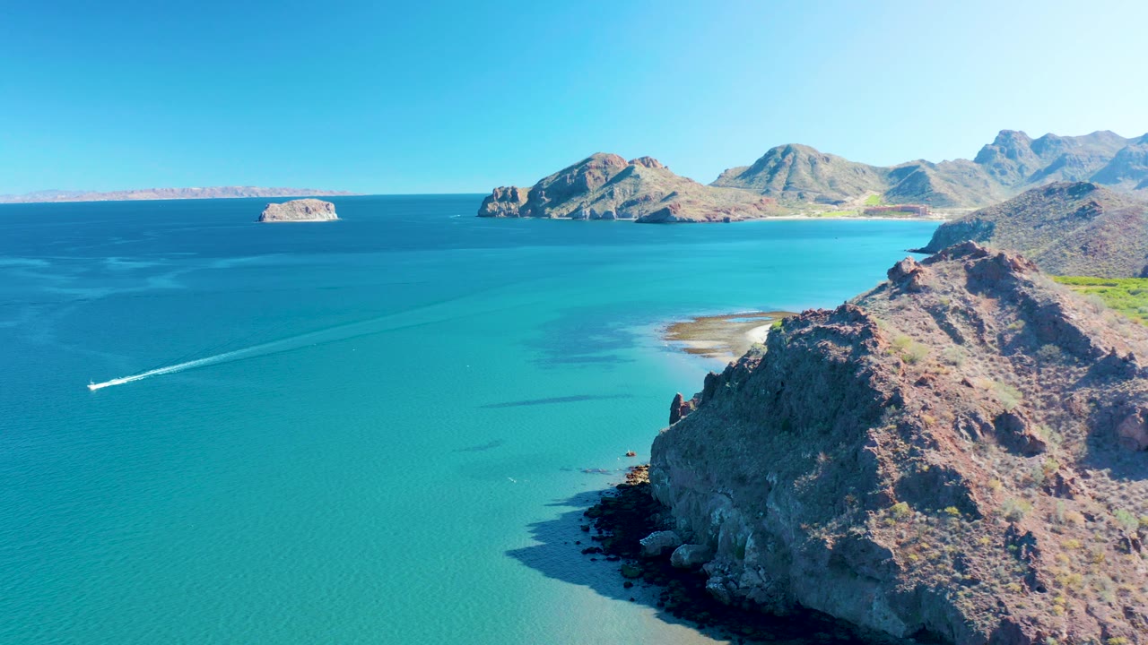 Wide aerial view over Ligui turquoise water with the boat wake near the left edge, rocky cliffs in the foreground, pale shallows by shore, and distant islands under a clear sky, no visible people.