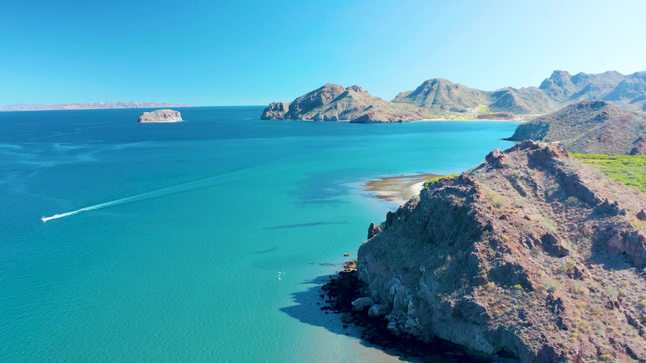 Aerial frame in Ligui with the boat and wake lower left, open turquoise bay across the center, rugged cliffs on the right, and distant rocky islands on the horizon, no visible people.