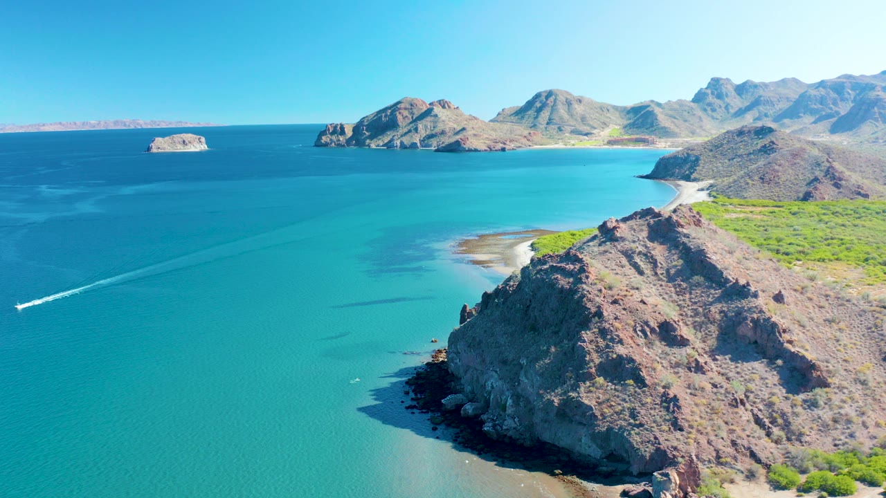 Aerial frame over Ligui, Baja California Sur showing turquoise water, a rocky foreground headland, distant mountains, offshore rocks, and no visible people.