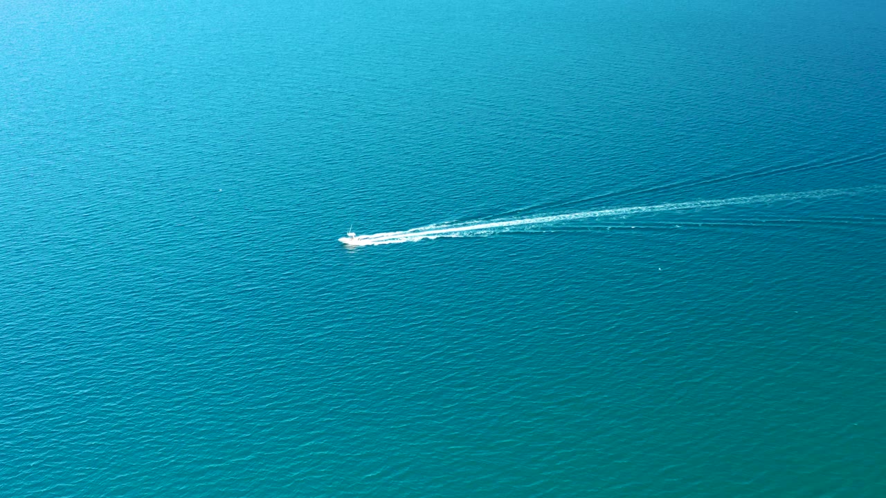 A small white boat rides near the upper part of the frame over deep blue and turquoise water in Ligui, Baja California Sur, with a bright foam trail and no visible people.
