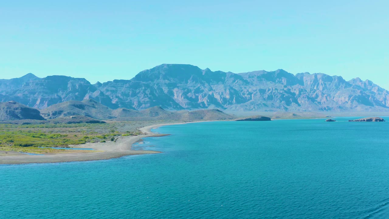 Aerial frame over Ligui, Baja California Sur, showing more open turquoise water, the sandy point along the left shore, distant small boats, rugged mountains, and no visible people.