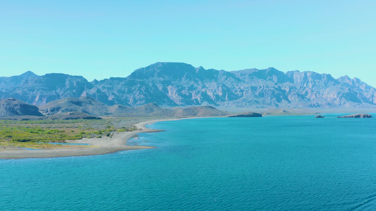 Beach curve and shallow turquoise water with mountains under clear sky