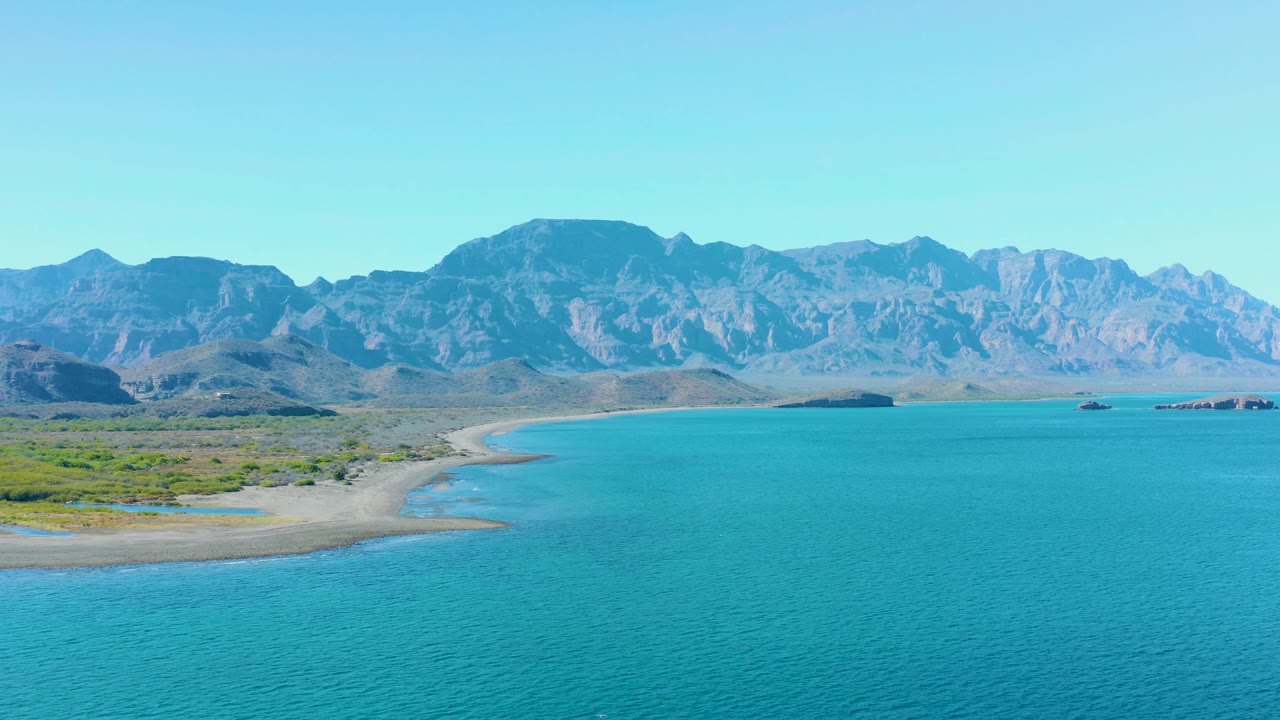 Wide aerial view of Ligui, Baja California Sur, with turquoise ocean in the foreground, a curving sandy shore at left, desert vegetation inland, distant mountains, and no visible people.