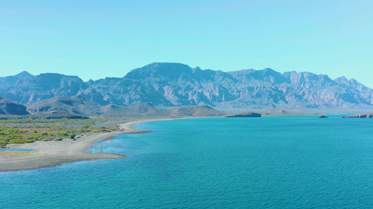 At 0:03, the left side of the Ligui shore shows more beach curve and scrubland, with turquoise water across the foreground and mountains beyond, no visible people.