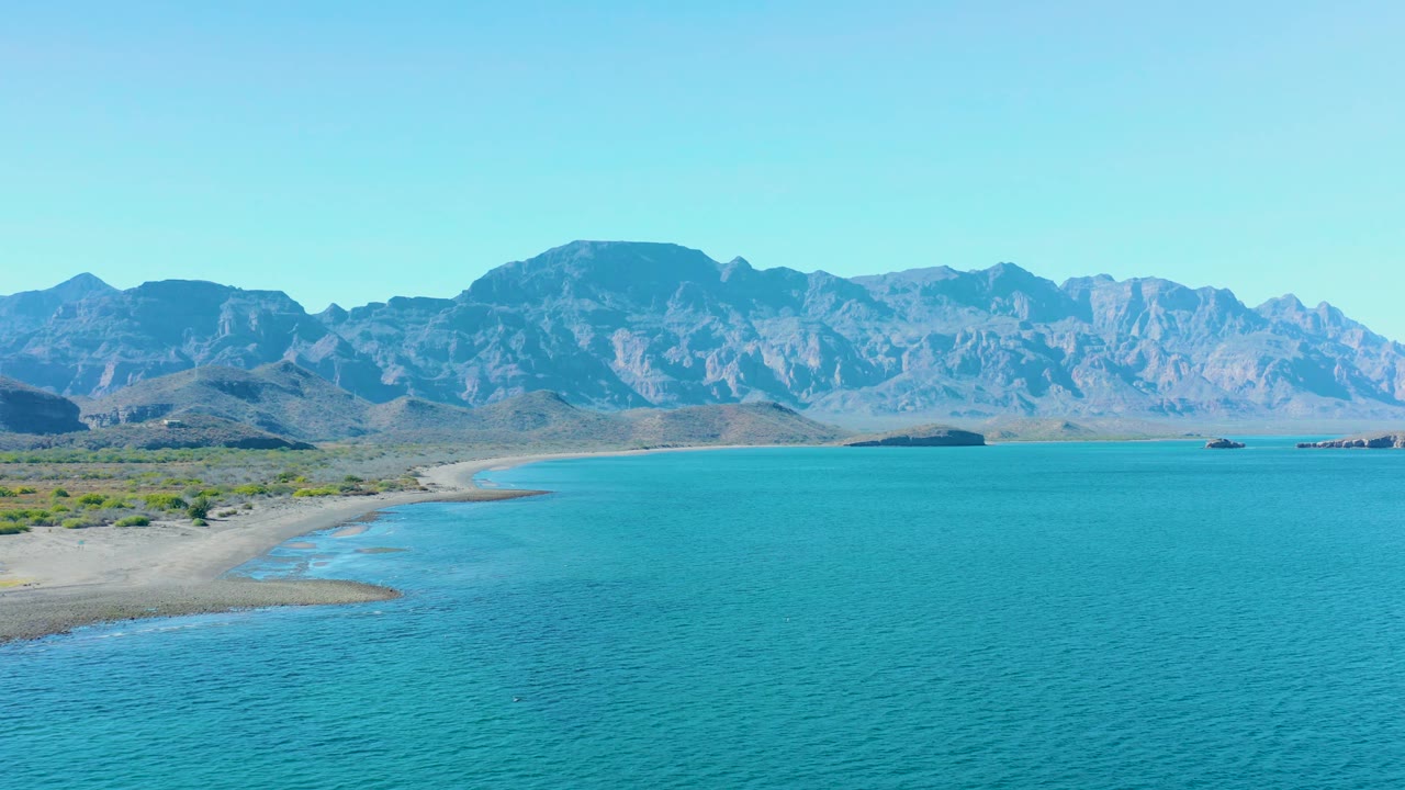 Wide coastal view of clear blue water, sand, and distant mountains.