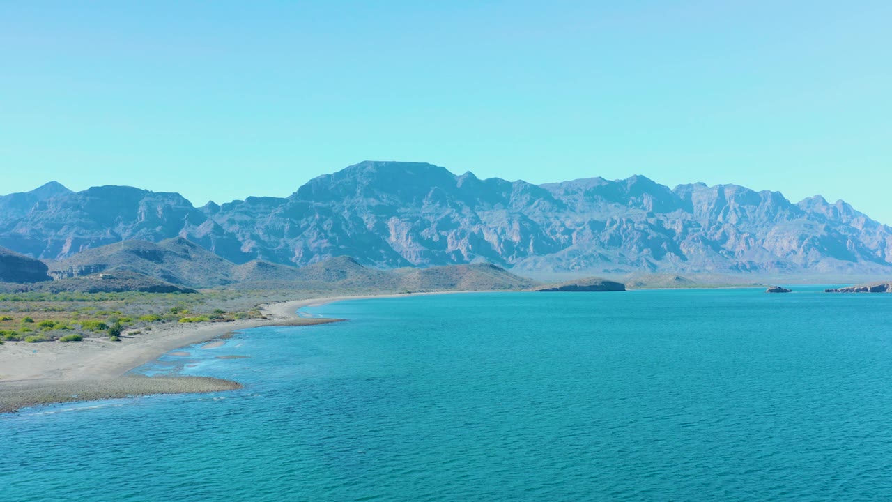 At 0:02 in Ligui, Baja California Sur, calm turquoise water dominates the foreground while a pale sandy beach curves along the left edge beneath a rugged mountain skyline, with no visible people.