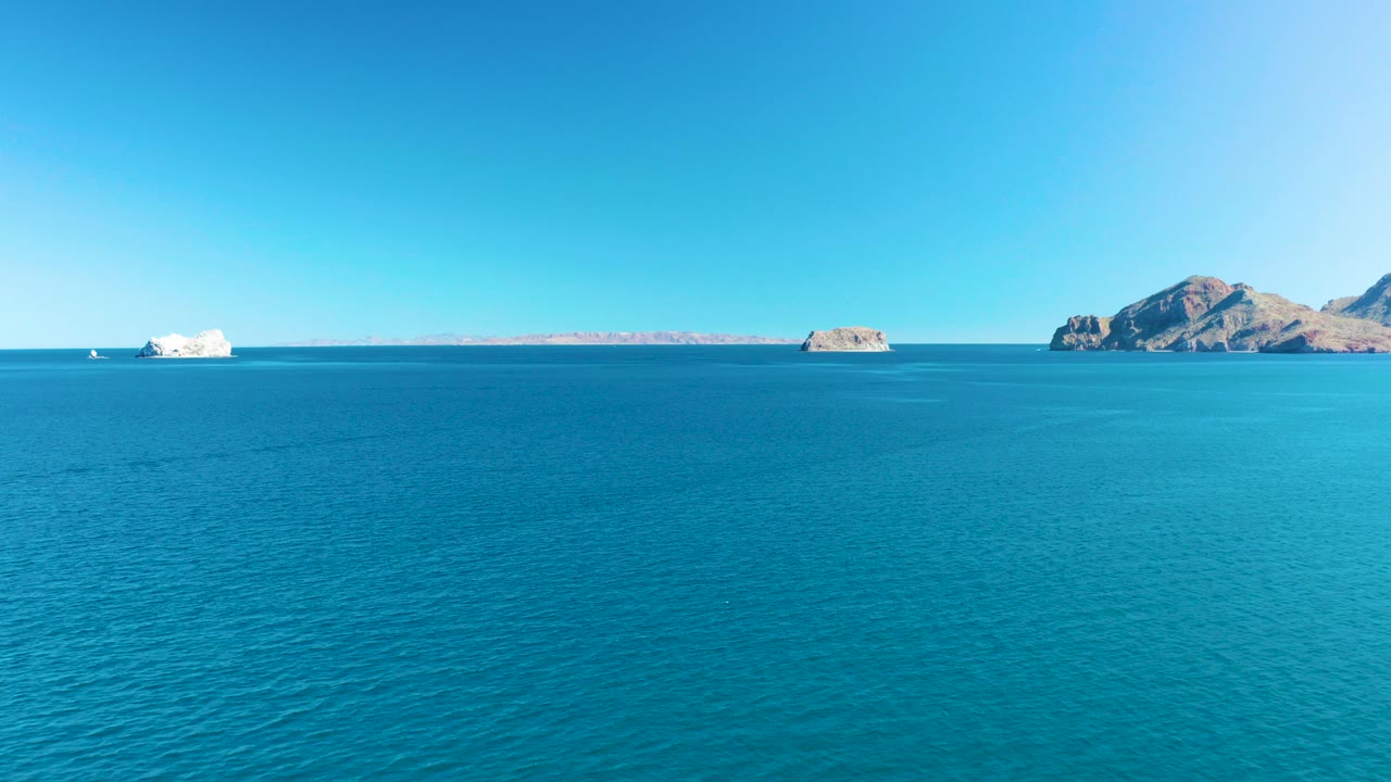 Wide view over turquoise water in Ligui, Baja California Sur, with distant rocky islands, a small white boat near the horizon, clear blue sky, and no visible people.