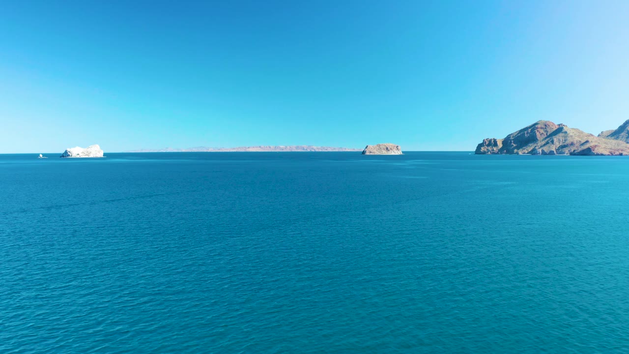 A clear horizon divides bright blue sky and turquoise water in Ligui, Baja California Sur, with rocky islands to the right and a small white boat farther left, no visible people.