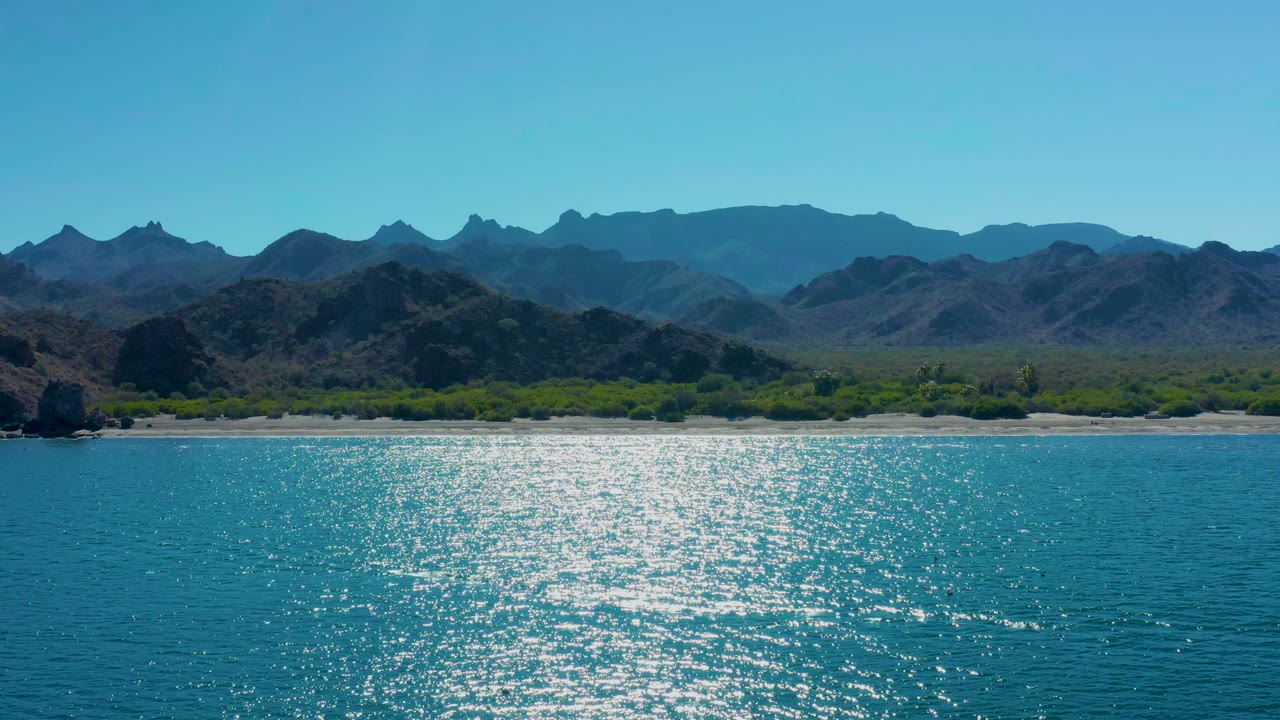 Turquoise ocean, sandy beach, green shoreline, and mountains under a clear sky.