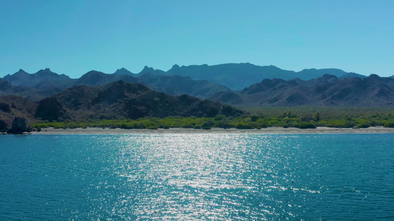 Aerial frame over Ligui, Baja California Sur showing bright sun glitter on the turquoise ocean, a sandy shoreline, green vegetation, rugged mountains, and no visible people.