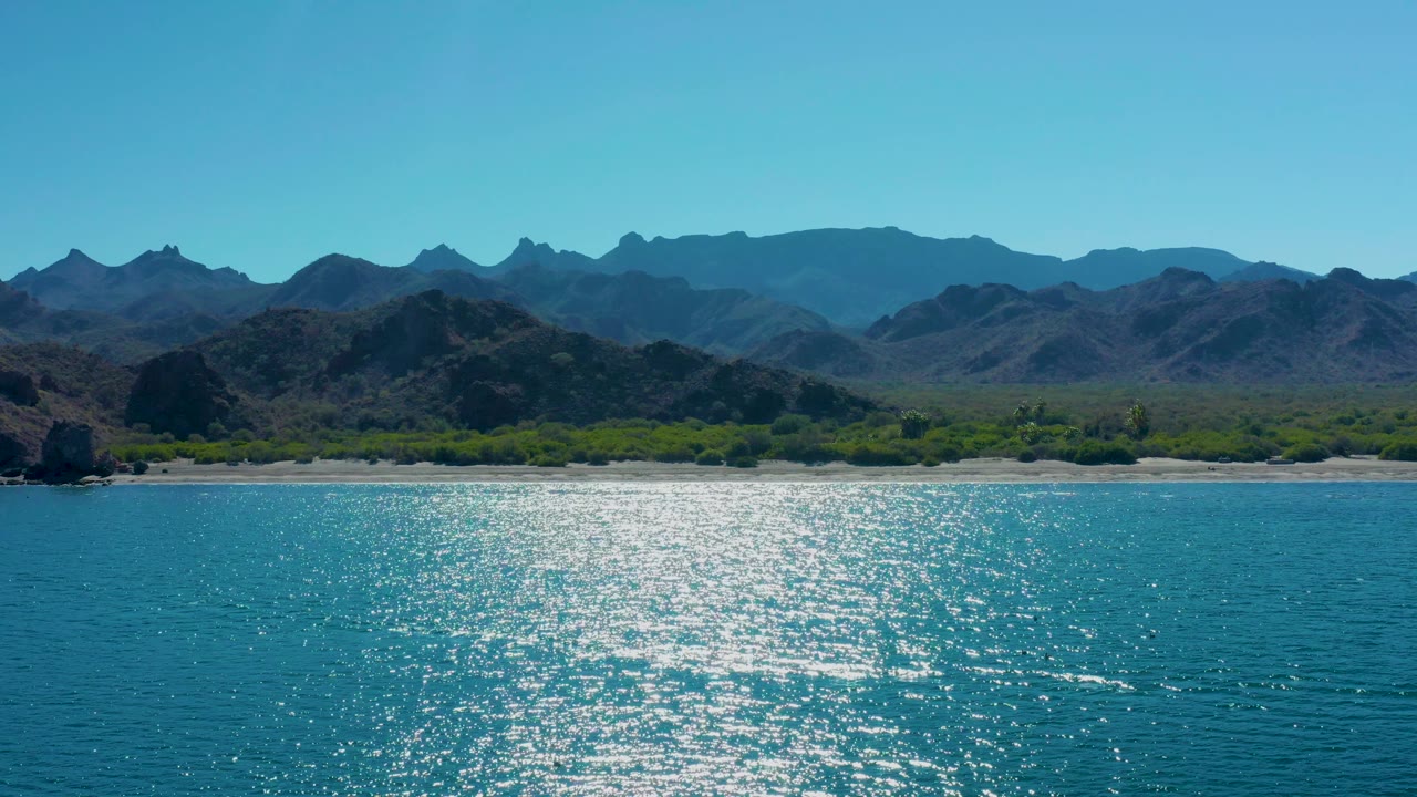 Wide aerial view of the Ligui coast with shimmering blue water in the foreground, pale sand and trees along shore, layered mountain ridges, and no visible people.
