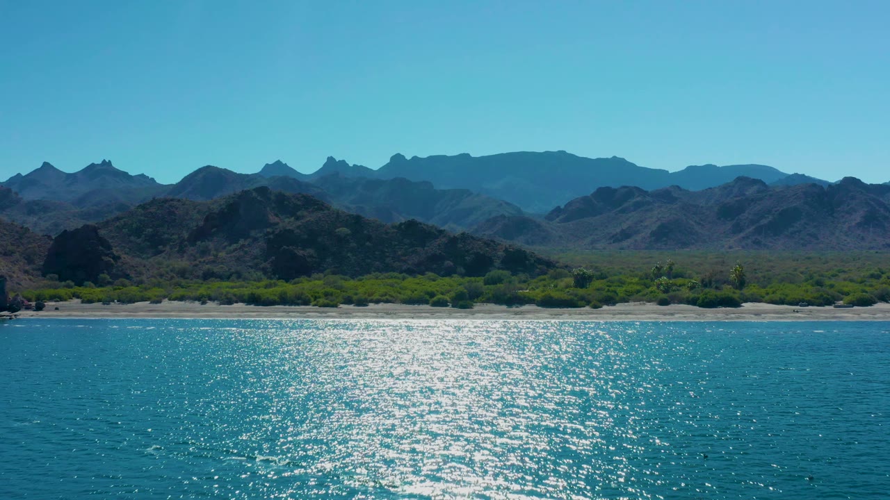 A second view along the Ligui coast shows a bright patch of reflected sunlight on the blue water, with a narrow beach, low greenery, mountains, and no visible people.