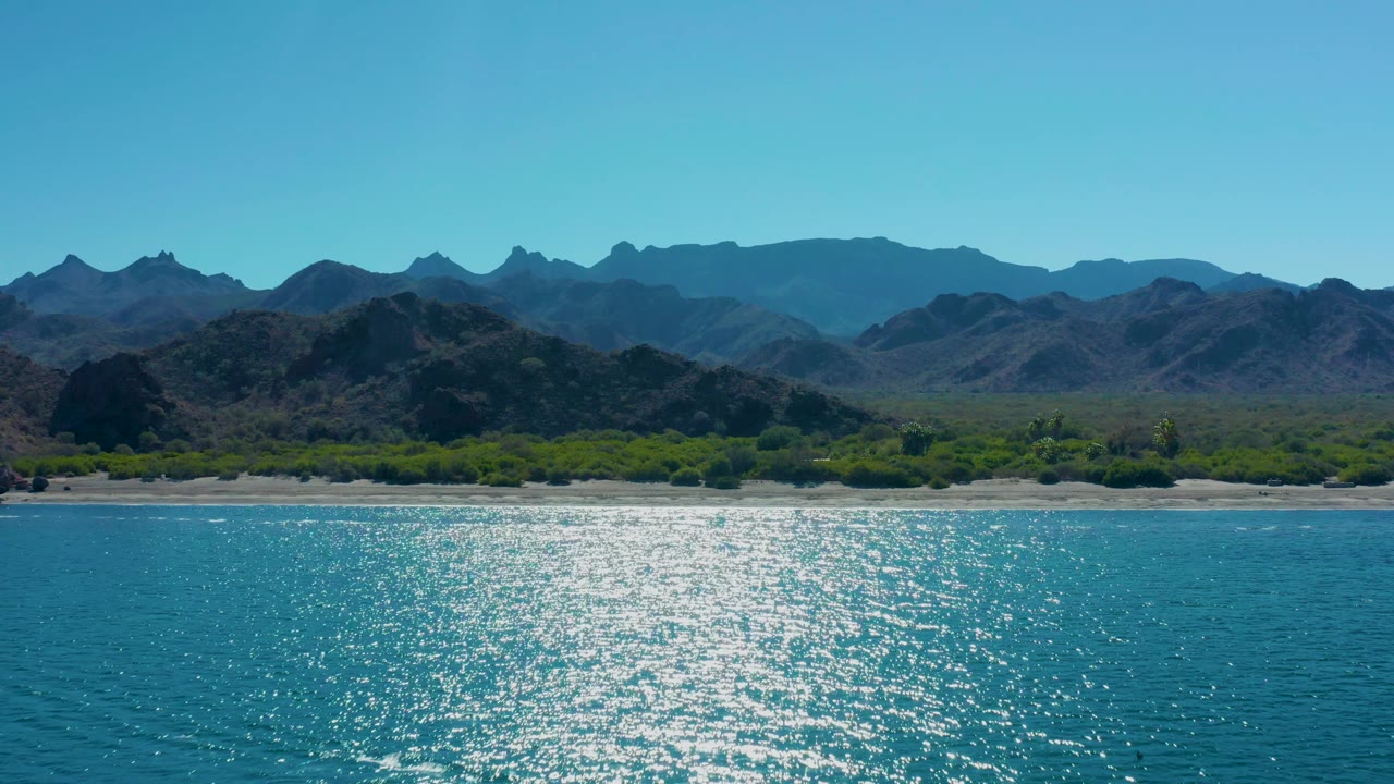 The Ligui shoreline remains centered with calm turquoise water catching hard sun, a thin pale sand band, sparse green vegetation, distant mountains, and no visible people.