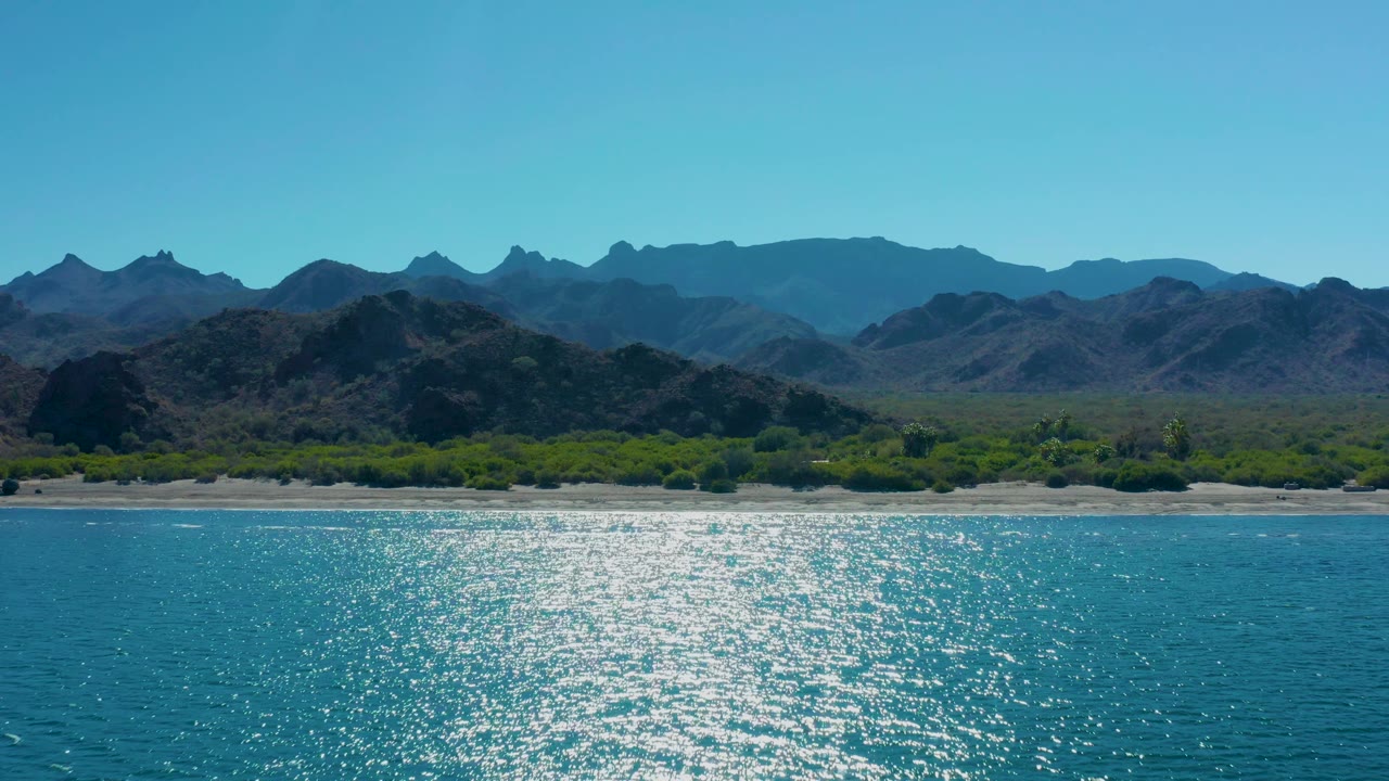 Mid-clip frame from Ligui, Baja California Sur with a broad blue ocean foreground, a bright reflection field, low green shore cover, jagged mountain silhouettes, and no visible people.