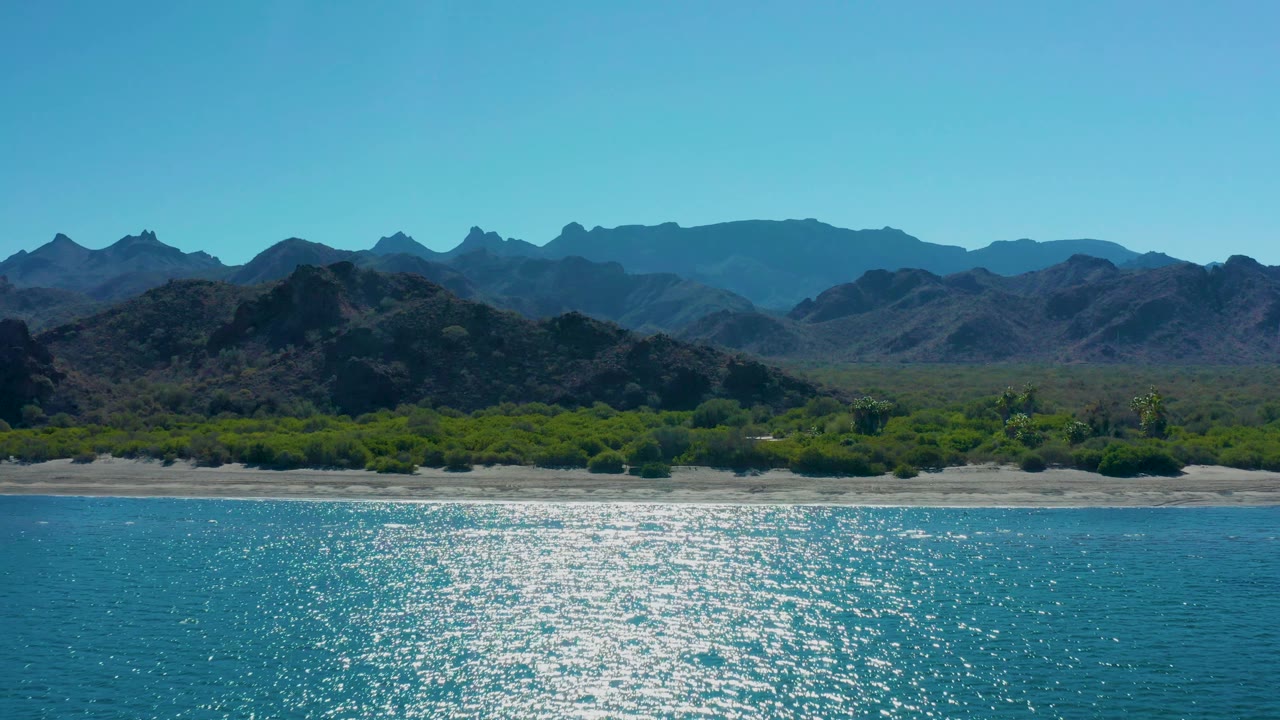 Aerial frame over Ligui coastline showing a broad patch of sunlight on the blue water, pale beach, dense green vegetation, layered mountain ridges, and no visible people.