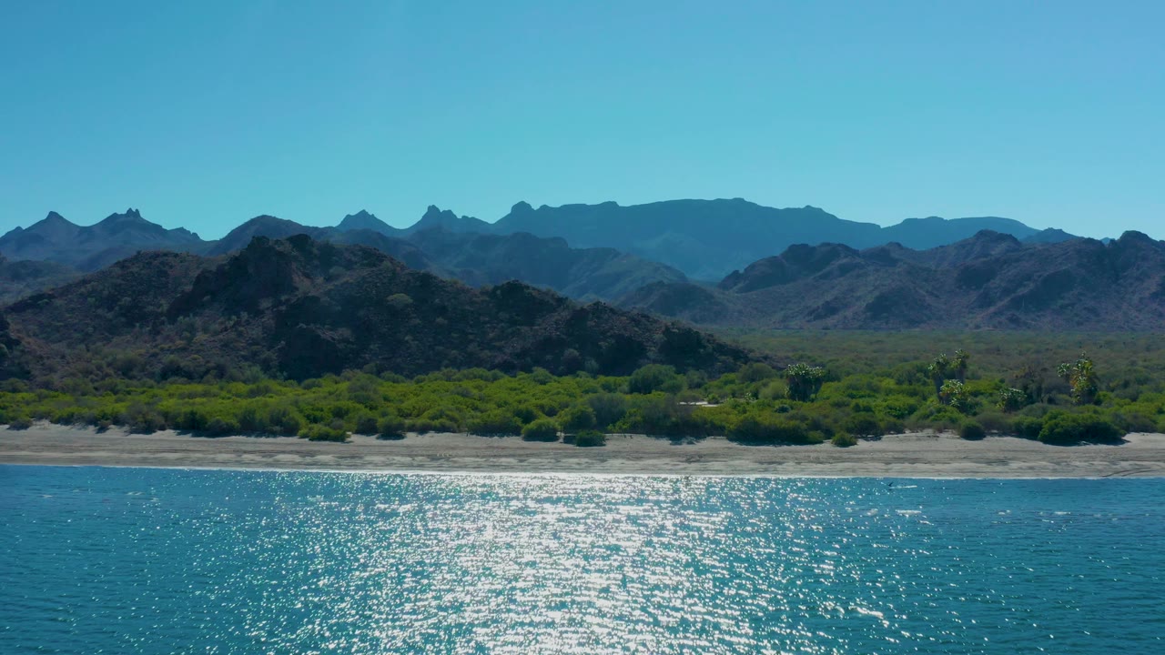 Closer shoreline view with turquoise water, sand, green vegetation, and mountains.
