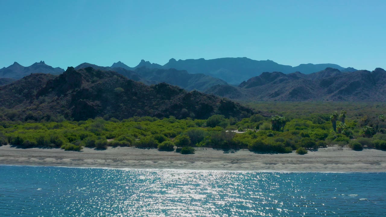 Wide aerial frame over Ligui, Baja California Sur, showing sunlit blue ocean in the foreground, pale sandy beach, green shoreline vegetation, rocky hills, distant mountains, and no visible people.