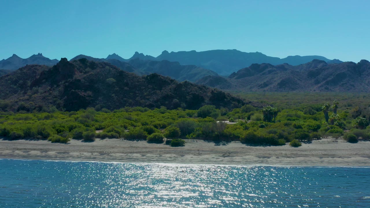 Drone-height frame over Ligui, Baja California Sur, showing blue ocean across the lower frame, a pale beach band, green coastal vegetation, a dark central hill, layered mountains, and no visible people.