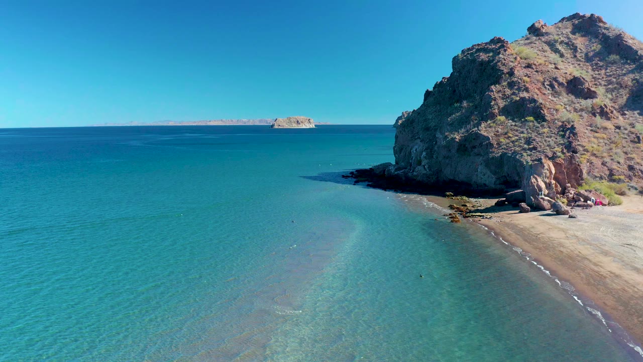 Aerial view in Ligui, Baja California Sur, with shallow turquoise water filling the foreground, a sandy beach at right, a rocky cliff, distant islands, and tiny people near the cliff base.