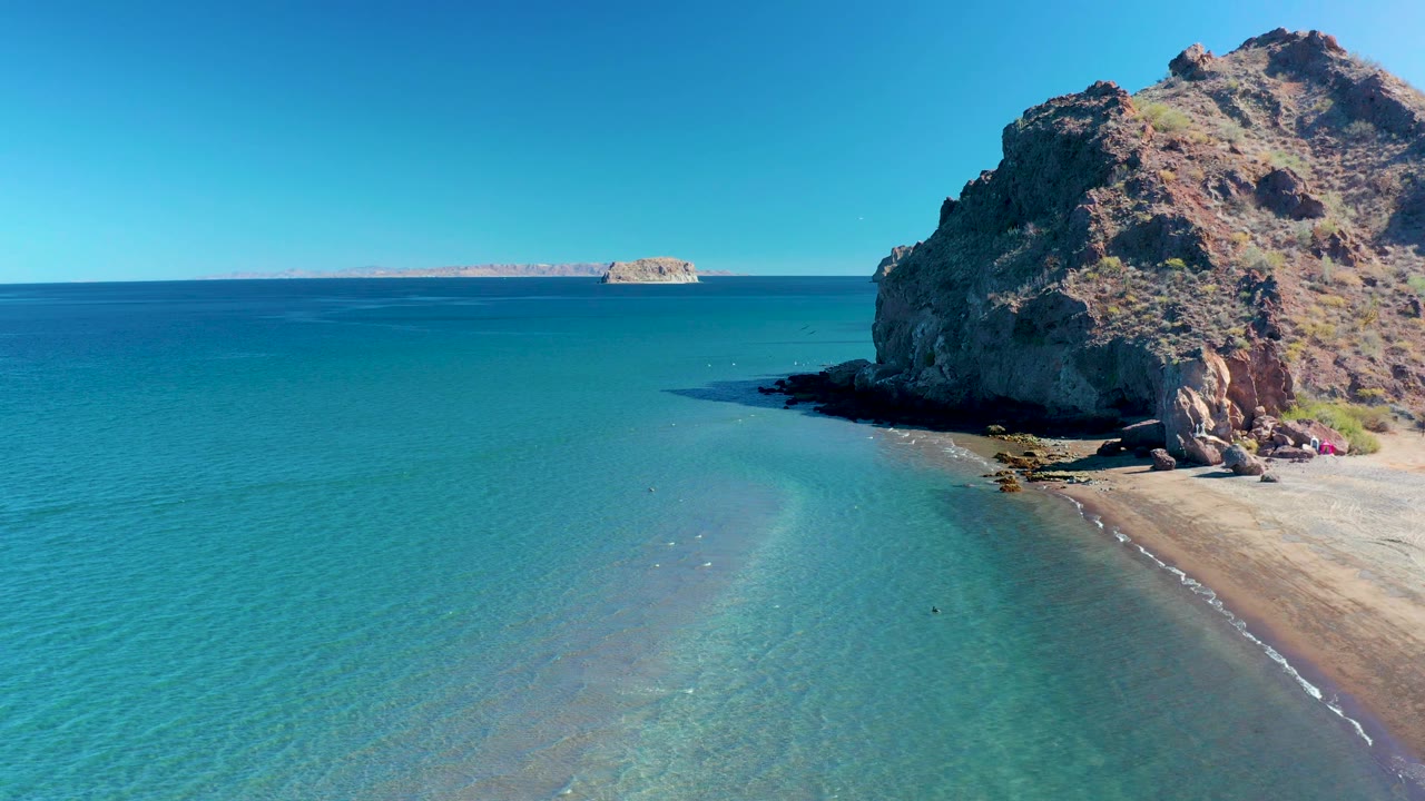 Ligui aerial frame with the rocky cliff cropped along the right edge, calm turquoise water across most of the frame, a narrow wash of waves on sand, distant islands, and tiny people by the cliff.