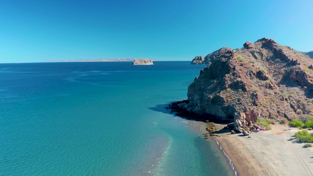 Wide aerial coastline with turquoise water, rocky cliff, sand, and distant islands.