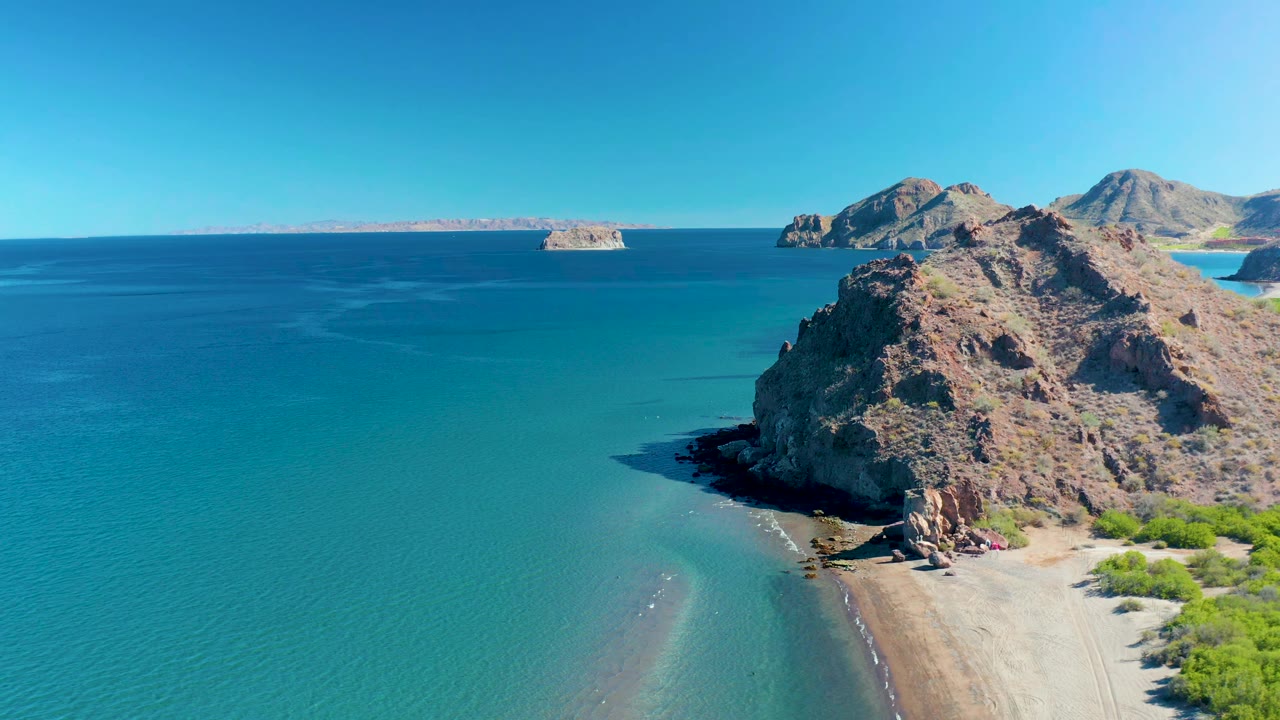 Aerial view in Ligui with the rocky point slightly farther right, shallow turquoise water curving into the sandy beach, distant landforms across the horizon, and no visible people.