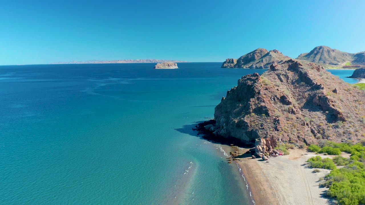 Aerial frame over Ligui showing the rocky headland dominating the right side, blue-green water across the left, a pale beach at the base, and no visible people.