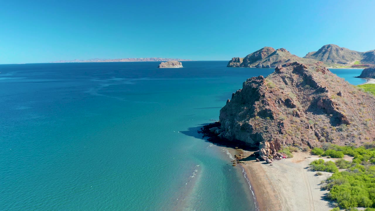 Aerial view of the Ligui coast with open turquoise water in the foreground, the rocky cliff and sandy beach on the right, distant land across the sea, and no visible people.