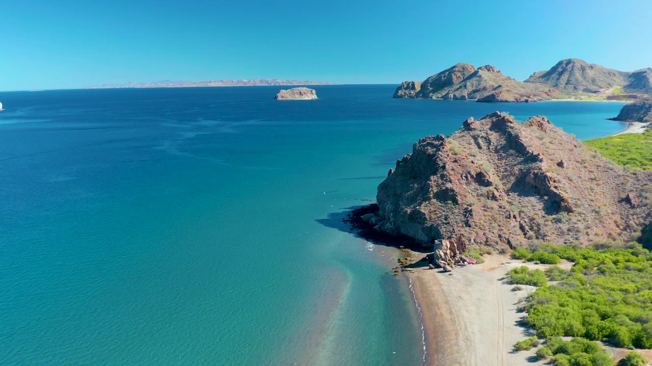 Aerial frame over Ligui, Baja California Sur shows turquoise water wrapping around a rocky headland, a pale beach, green shoreline vegetation, distant islands, and no visible people.